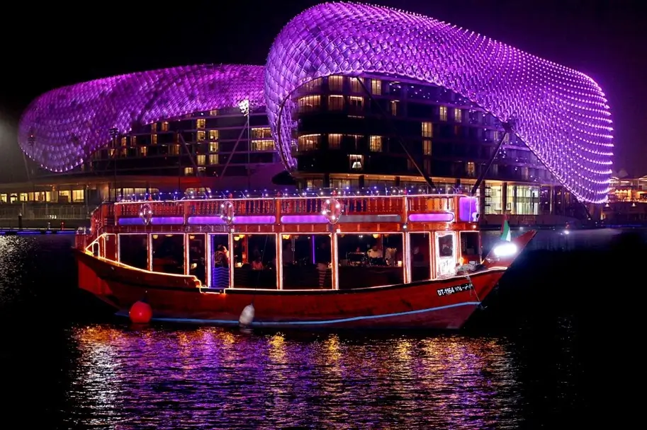 Wooden double-decker Abu Dhabi dhow lit in red and purple, moored at night facing the W Abu Dhabi