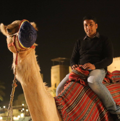 Seyed Shifaam riding a camel draped in a red patterned saddle blanket at night, with a fort tower behind