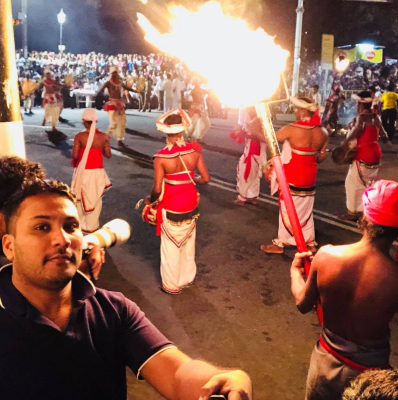 Seyed Shifaam at street level during the Kandy Esala Perahera with traditional dancers and fire performers behind him