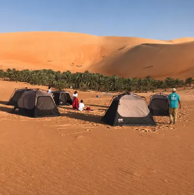 Arabiers overnight desert camping setup with tents beside a palm oasis and high Liwa dunes, managed by Mujeeb Rahman for desert guests