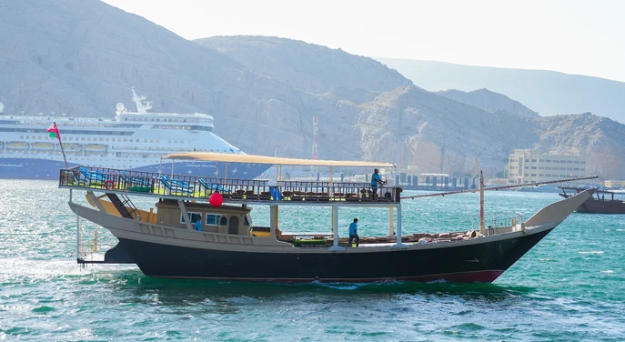 Traditional dhow sailing near Khasab harbour with a cruise ship and mountains behind